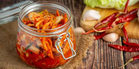 Słoik z kimchi na tle desek, obok kapusta, czosnek i papryczki chili.
