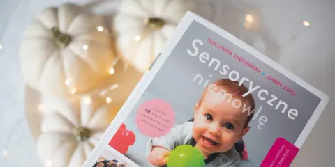 Okładka książki "Sensoryczne niemowlę" z uśmiechniętym dzieckiem, dyniami i lampkami.