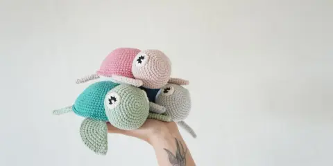 Una mano sostiene un trío de adorables tortugas a crochet: una verde menta, una rosa y una gris claro.