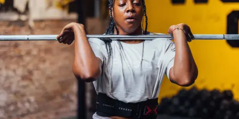 Frau mit geflochtenen Haaren hebt eine Hantel für Jumping Squats. Sie trägt einen Trainingsgürtel.