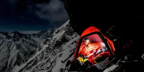 Nocny obóz na zboczu himalajskiego parbatu. W pomarańczowym namiocie rozświetlonym czołówką odpoczywa alpinista, otoczony surowym, ośnieżonym krajobrazem.