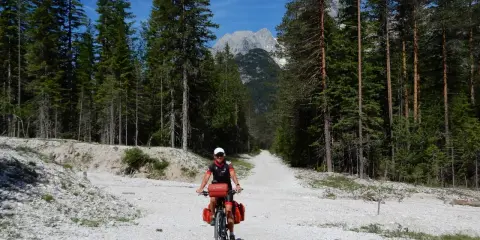 Ciclista su strada sterrata tra i pini, con le vette delle Dolomiti sullo sfondo. Un'avventura sui percorsi bici Dolomiti.