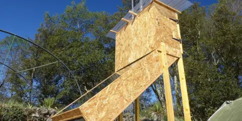 Un sechoir solaire artisanal en bois, avec un panneau incliné vitré, sous un ciel bleu.