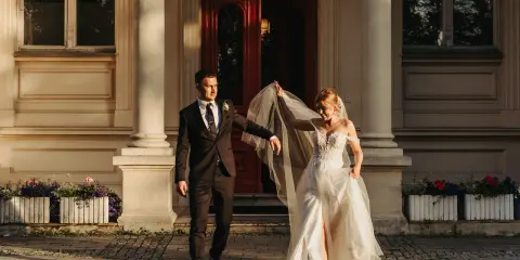 Para młoda podczas sesji zdjęciowej ślubnej schodzi po schodach przed eleganckim budynkiem. Panna młoda w zwiewnej sukni unosi welon, pan młody w garniturze patrzy na nią z uśmiechem.