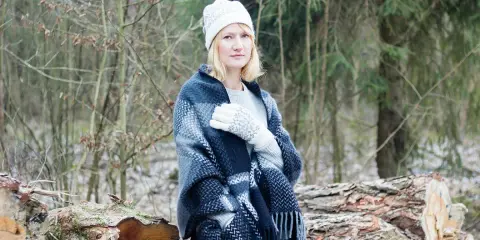Kobieta w czapce i rękawiczkach z wzorem, otulona grubym pledem. W tle las i ścięte drzewa. Ciepła wełna a bawełna w zimowej stylizacji.