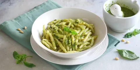 Un piatto di pasta con pesto fresco, guarnito con basilico e pinoli. Accanto, un mortaio con altro pesto.
