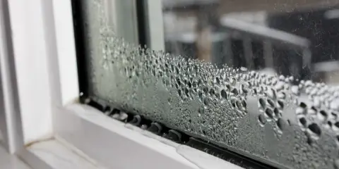 Goccioline d'acqua formano una **condensa interstiziale** sul vetro di una finestra, con un telaio bianco visibile.