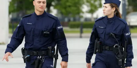 Dwóch policjantów w granatowych mundurach, z widocznymi stopniami w policji, idzie obok siebie.