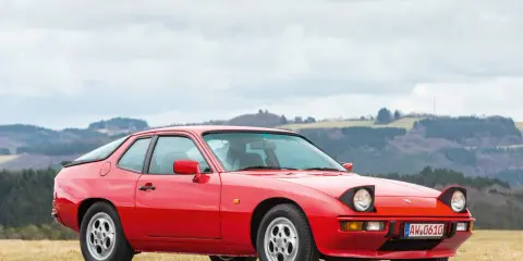 Roter Porsche 925 mit ausklappbaren Scheinwerfern steht auf einer Straße, im Hintergrund bewaldete Hügel.
