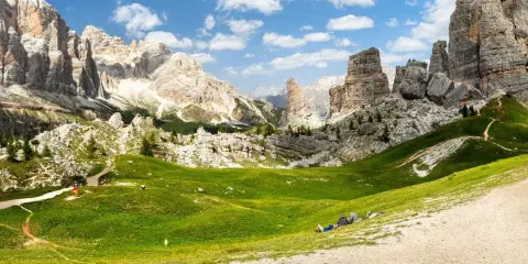 Un paesaggio mozzafiato nelle Dolomiti con le iconiche 5 torri che si ergono maestose. Un prato verde brillante invita al relax, con persone che riposano sotto il cielo azzurro.
