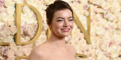 Emma Stone sorride, con un trucco che esalta i colori che stanno bene alle castane con pelle chiara. Lo sfondo è un muro di rose rosa e bianche.
