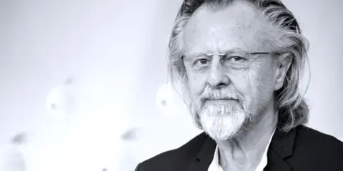 Portret mężczyzny z siwą brodą i długimi włosami, w okularach. Jeden z najlepsi współcześni kompozytorzy.