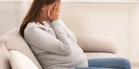 Kobieta w ciąży siedzi na kanapie, zakrywając twarz dłońmi. Jej postawa wyraża głęboki smutek, być może związany z diagnozą wady letalnej.