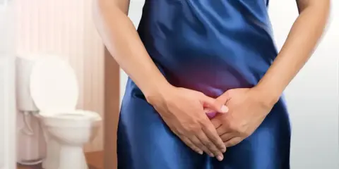 Kobieta w niebieskiej sukience odczuwa ból brzucha, być może spowodowany długim trzymaniem moczu. W tle widoczna jest toaleta.