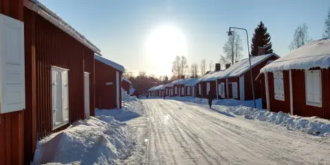 Vinterdag i kyrkbyn Gammelstad. Röda trähus med snö på taken kantar en snötäckt väg. Solen skiner starkt på en klarblå himmel.