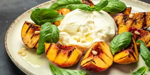 Burrata z grillowanymi brzoskwiniami, bazylią i orzeszkami piniowymi na talerzu.