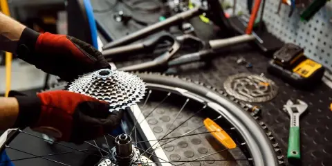 Ręce w rękawiczkach trzymają kasetę z zębatek, przygotowując rower do konserwacji. Na stole leżą narzędzia.