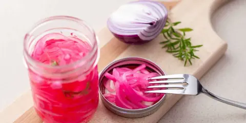 Pickles oignon rouge vibrants dans un bocal, avec une moitié d'oignon rouge et une branche de romarin sur une planche à découper.