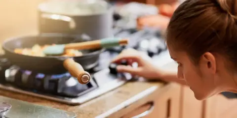 Mujer ajustando la perilla de la estufa de gas, con una sartén de verduras cocinándose. La llama azul indica que usa butano o gas natural.