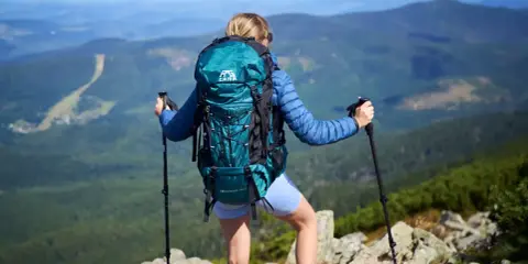 Kobieta z turkusowym plecakiem trekkingowym i kijkami na szczycie góry, podziwiająca panoramę.