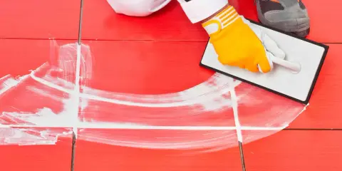 Man con guantes amarillos aplica lechada blanca a las juntas de baldosas rojas en el suelo exterior.