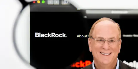 Uśmiechnięty mężczyzna w okularach, obok logo BlackRock. Larry Fink, jeden z założycieli BlackRock, na tle strony internetowej firmy.