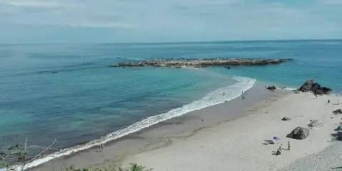 La **playa de Pechón** se extiende con arena dorada y aguas turquesas. Unas rocas emergen del mar, mientras algunas personas disfrutan del sol y el mar.