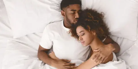 Ein Paar schläft eng zusammen in einem Bett. Er hält sie schützend im Arm, während sie sich an ihn kuschelt.