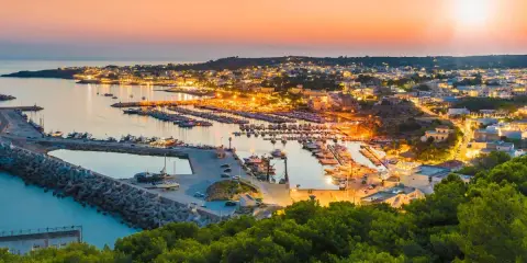 Porto illuminato al tramonto, un invito al turismo Salento. Barche ormeggiate e case che brillano sulla costa.