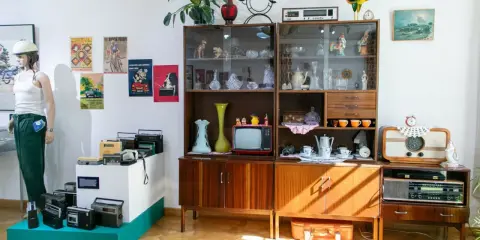 Stylizacja PRL: manekin w białej koszulce i zielonych spodniach, meblościanka z bibelotami, radia i telewizor.