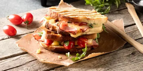 En staplad grilled cheese med skinka, tomater och ruccola på en träyta.