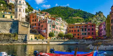 Colorate case a picco sul mare, barche rosse e blu in primo piano, un campanile svetta sulla collina. Un sogno di vacanze 5 Terre.