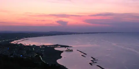 Panoramica Gabicce al tramonto, con cielo rosa e viola, mare calmo e costa illuminata.