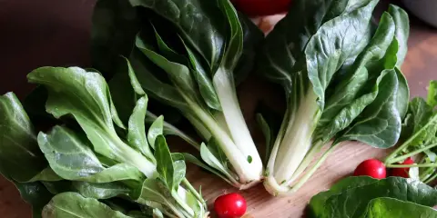 Färska bladgrönsaker, som mangold odla, ligger på en träskärbräda tillsammans med rädisor och en röd paprika.