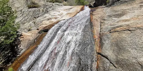 El agua desciende por la roca en la cascada del Hornillo, rodeada de vegetación y rocas graníticas bajo un cielo azul.