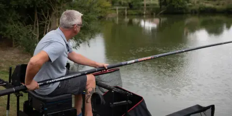 Mężczyzna siedzi nad wodą, przygotowując wędkę na grunt. Wędka jest długa, z widocznym napisem "CARPCOVER".