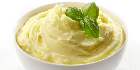 Una ciotola bianca piena di soffice crema di patate, guarnita con foglie di basilico fresco.