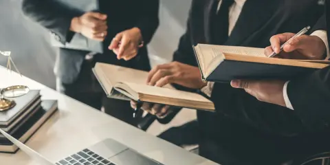 Trzy osoby w garniturach analizują dokumenty i książki przy biurku z laptopem i wagą sprawiedliwości.