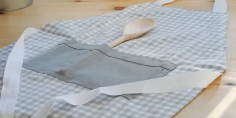 Gotowy fartuch kuchenny w kratkę, z drewnianą łyżką w kieszeni. Idealny poradnik, jak uszyć fartuch kuchenny.