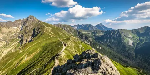 Widok na szlak turystyczny prowadzący przez zielone zbocza Tatr. W oddali majaczą kolejne pasma górskie, a niebo zdobią białe chmury. Kasprowy Wierch w całej okazałości.