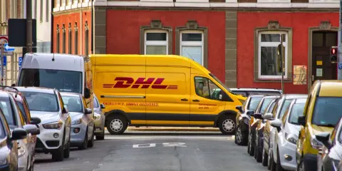 Żółta furgonetka DHL stoi zaparkowana na ulicy, obok innych samochodów. To miejsce, do którego pracują kurierzy DHL, by dostarczać paczki.