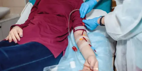 Pacjentka leży, a pielęgniarka w rękawiczkach podłącza przetokę do dializ. Krew płynie przez rurki.