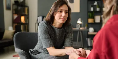 Kobieta w szarej sukience siedzi w fotelu, trzymając dłonie drugiej osoby. To scena wspierającej terapii gestalt.