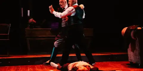 Dwóch mężczyzn tańczy tango na scenie, trzeci leży na podłodze.