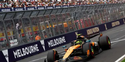 Un coche de Fórmula 1 naranja y negro acelera en Melbourne. Los aficionados vitorean. ¡Qué ganas de que empiece la Fórmula 1 2026!