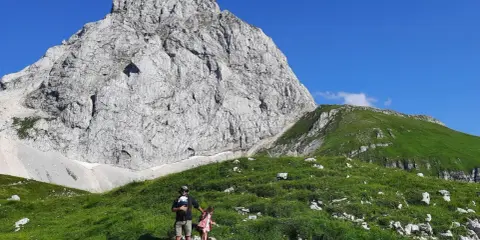 Ojciec z dzieckiem na rękach i córką obok na zielonym zboczu. W tle potężna, skalista góra, idealna na pierwsze górskie wędrówki.