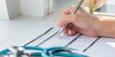 Dłoń pisząca długopisem na formularzu medycznym, obok stetoskopu.