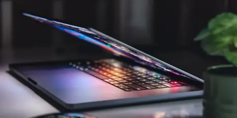 Otwarty notebook z podświetlaną klawiaturą i ekranem, obok myszka i roślina. To jest właśnie notebook, narzędzie do pracy i rozrywki.