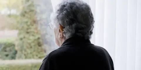 Starsza pani z siwymi włosami patrzy przez okno. Czeka na emeryturę po mężu, wspominając wspólne chwile.