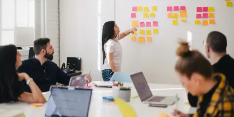 Une femme explique des techniques d'animation de réunion participative devant un mur couvert de post-its.
